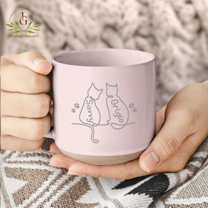 May include: A pale pink ceramic mug with a light brown base. The mug features a line drawing of two cats with the names "Savvy" and "Grigio" and paw prints. The mug is held by a person.