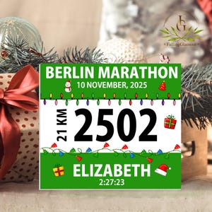 May include: A square Berlin Marathon race bib featuring a green and white design with the text BERLIN MARATHON, 10 NOVEMBER, 2025, 21 KM, 2502, ELIZABETH, and 2:27:23. The bib is decorated with Christmas lights, a snowman, gifts, and a Santa hat.