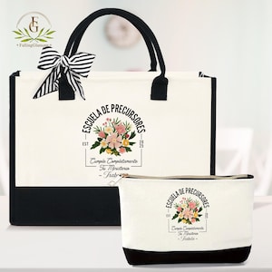 May include: A white tote bag and matching cosmetic bag with black accents. Both bags feature a floral design and the text "ESCUELA DE PRECURSORES". The tote bag has black handles and a striped bow.