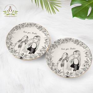 May include: Two white ceramic plates with a gold rim. Each plate features a black and white illustration of three women and the text "Love you Bestie". The plates are surrounded by a floral border.