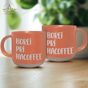 May include: Two orange ceramic coffee mugs with the words "BOREI PRI HACOFFEE" in white. The mugs have a grey base and a curved handle. The mugs are on a wooden surface.
