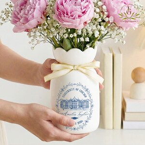 Pu&ograve; includere: Un vaso in ceramica bianca con bordo smerlato, legato con un nastro dorato, contiene peonie rosa e gypsophila bianca. Il vaso presenta un disegno floreale blu con "UNIVERSITY OF CHICAGO" e un nome e anno di laurea.