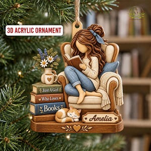 May include: A 3D acrylic ornament featuring a person with brown hair sitting in a chair reading a book. Beside them is a stack of three books labeled Just A Girl Who Loves Books, a vase with flowers, a mug, and a sleeping orange cat. The name Amelia is on a plaque.