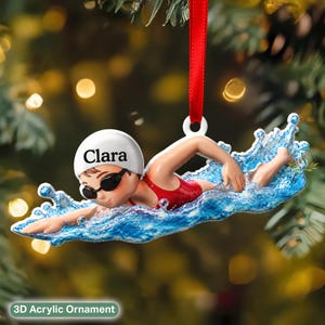 May include: A 3D acrylic ornament featuring a swimmer in a red swimsuit and white cap with the name Clara printed in black. The swimmer is depicted in a freestyle stroke surrounded by a blue water splash effect, suspended by a red ribbon.