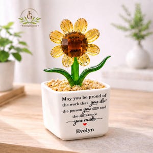 May include: A glass sunflower figurine with yellow petals and a brown centre, set in a white square ceramic pot filled with small pebbles. The pot features black printed text: May you be proud of the work that you do, the person you are and the difference you make, followed by the name Evelyn and a small red heart.