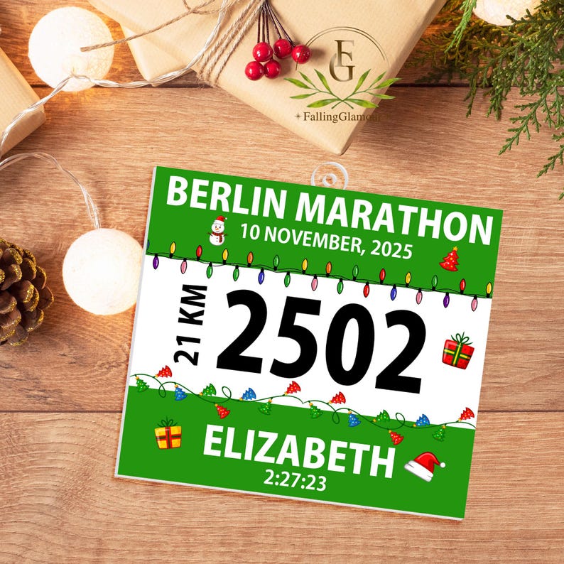 May include: A square Berlin Marathon race bib ornament featuring a green and white design with the text BERLIN MARATHON, 10 NOVEMBER, 2025, 21 KM, 2502, ELIZABETH, and 2:27:23. It includes illustrations of Christmas lights, a snowman, and gifts.