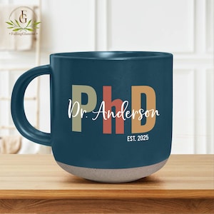 May include: A dark blue ceramic mug with a white inscription that reads "PhD Dr. Anderson EST. 2025". The letters "PhD" are in colorful blocks, with "P" in beige, "h" in pink, and "D" in orange.
