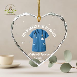 K&ouml;nnte beinhalten: Herzf&ouml;rmiger Kristallschmuck mit goldener Kette. Das Ornament zeigt eine blaue Krankenschwesteruniform mit Stethoskop und den Text "OFFICIALLY DISCHARGED" und "Retired Nurse 2026".