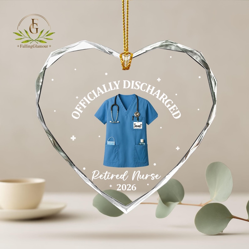 K&ouml;nnte beinhalten: Herzf&ouml;rmiger Kristallschmuck mit goldener Kette. Das Ornament zeigt eine blaue Krankenschwesteruniform mit Stethoskop und den Text "OFFICIALLY DISCHARGED" und "Retired Nurse 2026".