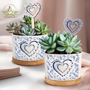 May include: Two white ceramic planters with blue floral patterns and the word "Grandma". Each planter contains a succulent and rests on a wooden base. Heart-shaped toppers with names are placed in the plants.