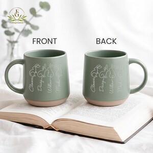May include: Two ceramic mugs with a sage green upper and tan base are displayed. Each mug features a white line drawing of four rabbits and the names Clover, Daisy, Willow, and Mocha. The mugs are on an open book.