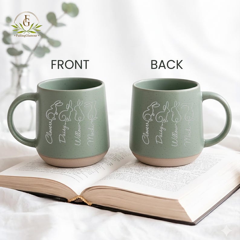 May include: Two ceramic mugs with a sage green upper and tan base are displayed. Each mug features a white line drawing of four rabbits and the names Clover, Daisy, Willow, and Mocha. The mugs are on an open book.