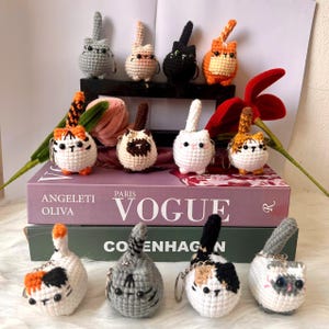 May include: A collection of handmade crochet cat keychains in various colors, including gray, black, orange, and white. The keychains are arranged on a shelf and on top of books with the words "Vogue" and "Copenhagen" visible.