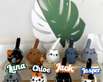May include: A collection of handmade crochet cat keychains in various colors, including black, brown, white, gray, and blue. Each keychain features a unique design and is labeled with a name. The keychains are displayed on a wooden shelf.