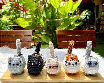 May include: Five handmade crochet cat keychains in a row. The cats are in various colors: gray, black, cream, white with orange stripes, and white with blue eyes. Each cat has a tail and a silver keychain ring. They are displayed on a wooden tray.