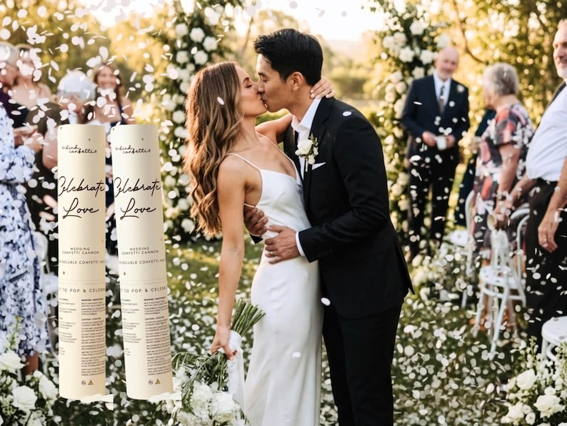 May include: A wedding celebration with a couple kissing, surrounded by falling confetti. Two confetti cannons are visible, with the words "Celebrate Love." The bride wears a white dress, and the groom is in a black suit.