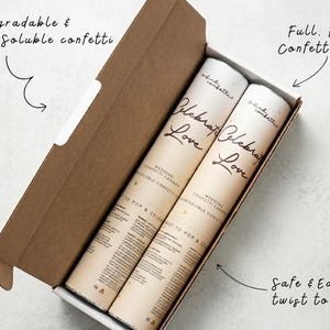 Op de afbeelding: Een bruine kartonnen doos met twee bruiloftconfettikanonnen. De kanonnen zijn cilindrisch, met de woorden "Celebrate Love" erop gedrukt. De doos bevat ook de tekst "Biologisch afbreekbare & in water oplosbare confetti".