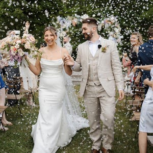 Op de afbeelding: Een pasgetrouwd stel loopt door een confetti regen. De bruid draagt een witte trouwjurk met spaghettibandjes en een boeket. De bruidegom draagt een beige pak. Gasten vieren de bruiloft.