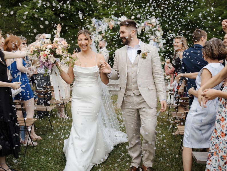 Op de afbeelding: Een pasgetrouwd stel loopt door een confetti regen. De bruid draagt een witte trouwjurk met spaghettibandjes en een boeket. De bruidegom draagt een beige pak. Gasten vieren de bruiloft.