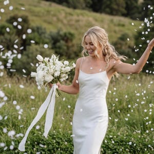 Op de afbeelding: Een vrouw in een witte jurk met spaghettibandjes lacht en gooit confetti in een veld. Ze houdt een boeket witte bloemen met lange witte linten vast. De achtergrond toont groen gras en bomen.