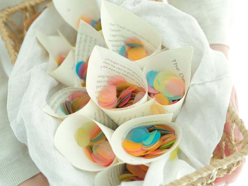 K&ouml;nnte beinhalten: Ein Korb gef&uuml;llt mit Papierkegeln, die buntes Konfetti enthalten. Die Kegel sind aus Buchseiten mit sichtbarem Text gefertigt. Der Text auf einem Kegel lautet "The Delicate."