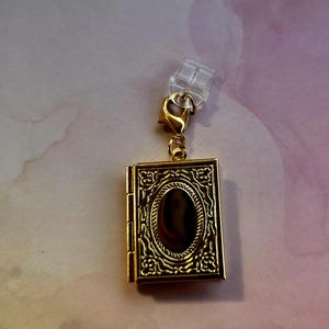May include: A gold-colored, rectangular charm in the shape of a book. The charm has an oval-shaped center and is decorated with floral and scroll designs. It is attached to a clear plastic clip with a gold-colored clasp.