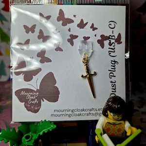 May include: A gold-colored USB-C dust plug in the shape of a small sword. The plug is attached to a clear plastic piece. The product is displayed on a card with butterfly graphics and the text "Mourning Cloak Crafts".
