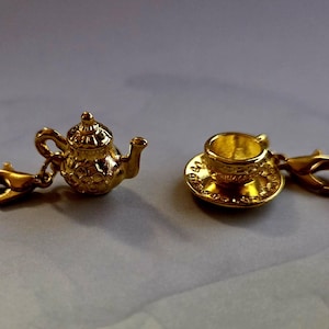 May include: Two gold-colored charms: a teapot and a teacup with saucer. The teapot charm has a detailed design, including a lid and spout. The teacup charm sits on a saucer. Both charms have gold-colored clasps and clear plastic attachments.