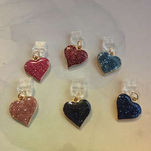 Puede incluir: Seis colgantes en forma de corazón en varios colores, incluyendo rosa, rojo, azul y negro, con superficies brillantes y borde dorado. Cada colgante está unido a un clip transparente. Los colgantes son probablemente para joyería o proyectos de manualidades.