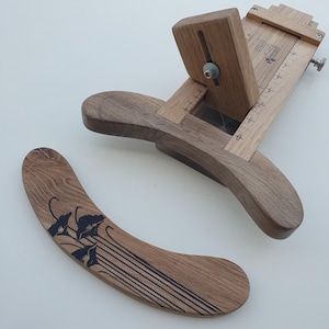 Pode incluir: Uma ferramenta de artesanato em couro de madeira com uma alça curva e uma lâmina. A ferramenta tem um acabamento castanho claro e um botão de ajuste de metal. Uma peça de madeira separada com um design floral também está presente.