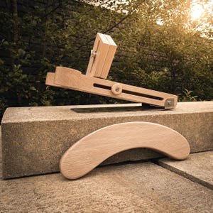 Op de afbeelding: Een verstelbaar gereedschap van lichtgekleurd hout met een lange horizontale arm met een ronde knop, een verticaal blokonderdeel en een apart gebogen houten stuk. Het mechanisme bevat een platte metalen basisplaat en zilverkleurige hardware.