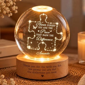 May include: A spherical glass night light featuring an etched puzzle piece design with the text "Never forget you are the piece that made the difference" and the name "Amanda". The light sits on a cylindrical wooden base with engraved text.