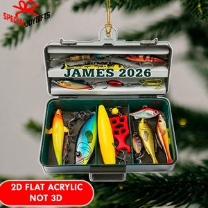 May include: A flat acrylic Christmas tree ornament shaped like an open fishing tackle box. The interior displays various colorful fishing lures and hooks. The text JAMES 2026 is printed in dark green across the center. A gold string is attached to the top.