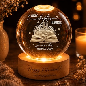 May include: A spherical glass night light on a cylindrical wooden base. The glass features an etched open book with wildflowers, butterflies, and the text A NEW CHAPTER BEGINS, Amanda, and RETIRED 2026. The wooden base is engraved with Happy Retirement.