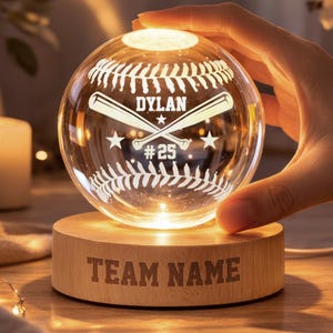 May include: A baseball-themed night light with a clear glass baseball design. The baseball has white stitching, crossed bats, the name "DYLAN", and the number "25". It sits on a wooden base with the text "TEAM NAME".