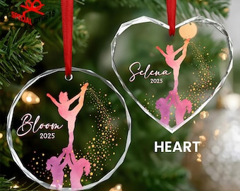 May include: Two clear glass ornaments with red ribbons hang from a Christmas tree. One is round with the word "Bloom" and the year "2025". The other is heart-shaped with the name "Selena" and the year "2025". Both ornaments feature a cheerleader design.