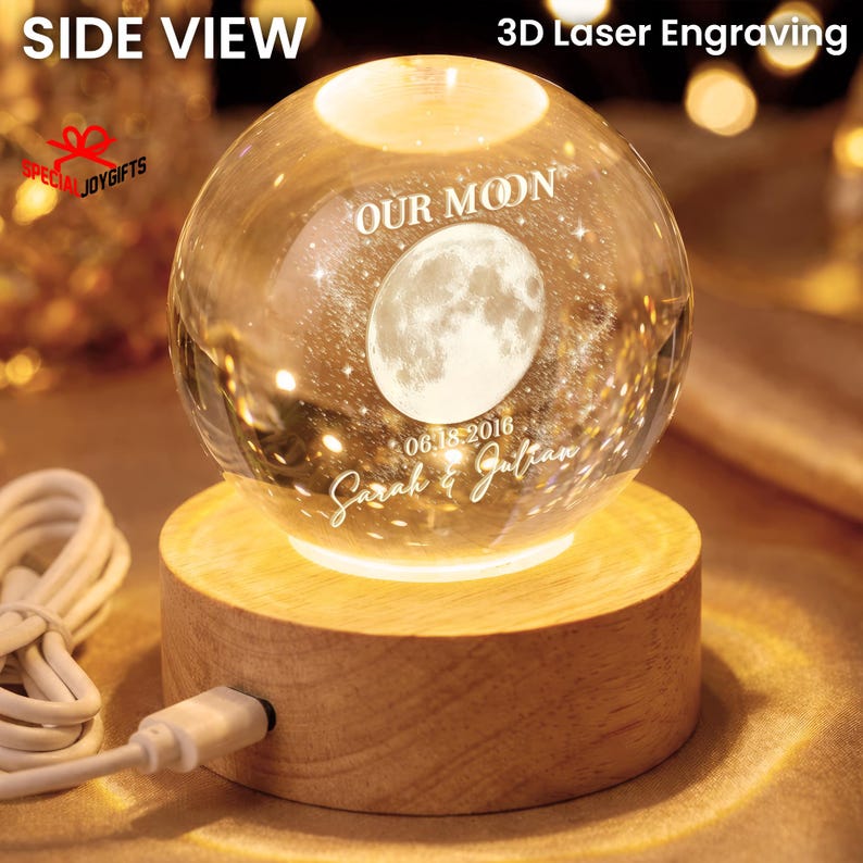 May include: A clear glass sphere featuring a 3D laser engraving of the moon, the text "OUR MOON", the date "06.18.2016", and the names "Sarah & Julian". The sphere rests on a round, light-colored wooden base with an attached white power cord.