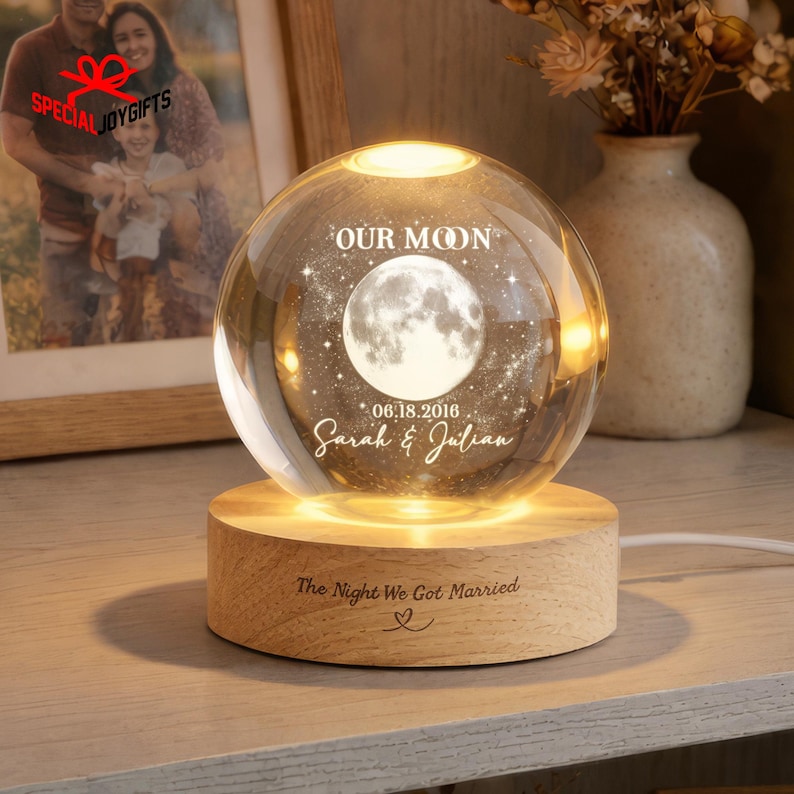 May include: A spherical glass night light featuring an internal laser-etched moon graphic with the text OUR MOON, 06.18.2016, and Sarah & Julian. The sphere rests on a circular wooden base engraved with The Night We Got Married and a heart icon.