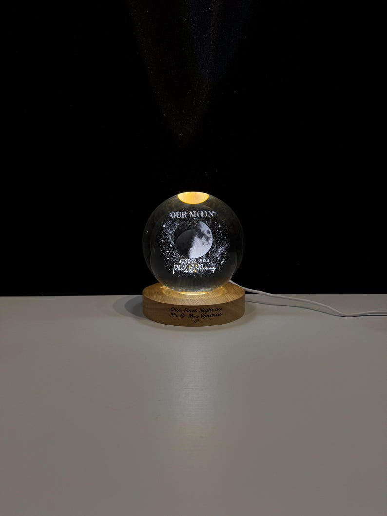 May include: A clear glass sphere night light featuring an etched moon graphic, the text "OUR MOON", "JUNE 22, 2028", and "Phil & Penny". The sphere rests on a circular wooden base engraved with the text "Our First Night as Mr & Mrs Vondras".