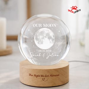 May include: A clear glass sphere featuring an etched moon and star design with the text "OUR MOON", "06.18.2016", and "Sarah & Julian". The sphere rests on a cylindrical light-colored wooden base engraved with "The Night We Got Married" and a heart.