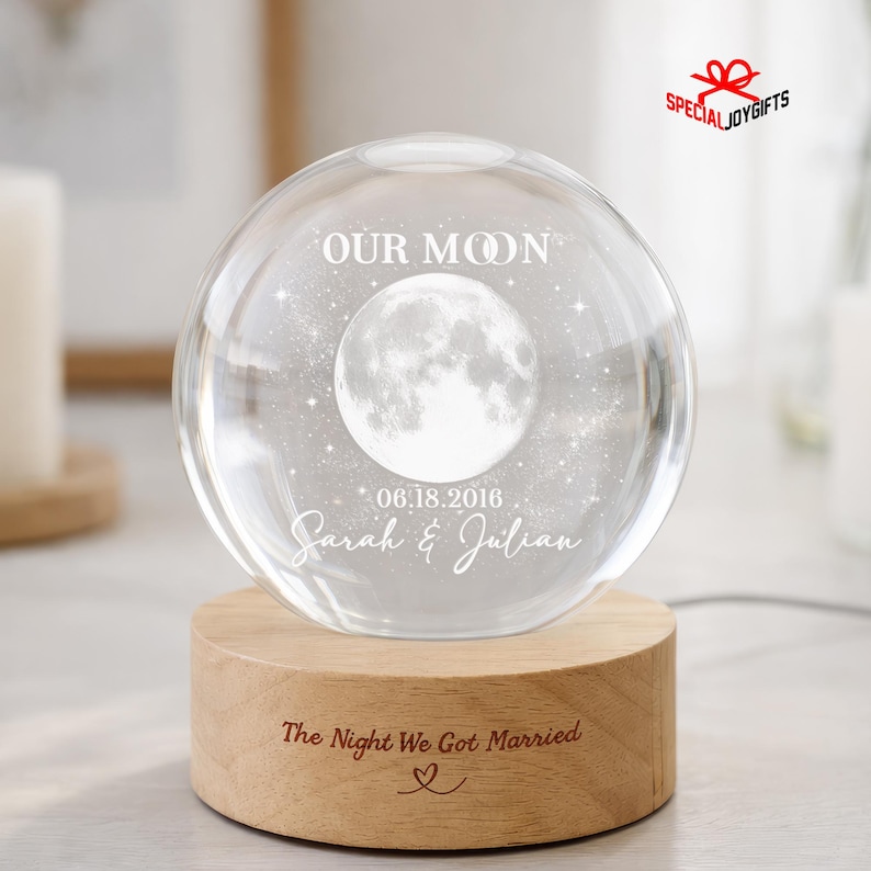 May include: A clear glass sphere featuring an etched moon and star design with the text "OUR MOON", "06.18.2016", and "Sarah & Julian". The sphere rests on a cylindrical light-colored wooden base engraved with "The Night We Got Married" and a heart.