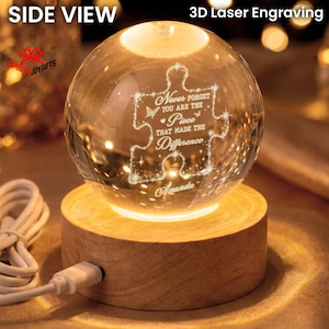 May include: A clear glass sphere with a 3D laser-engraved puzzle piece design featuring the text "Never forget you are the piece that made the difference" and the name "Amanda," resting on a round, light-colored wooden base with a white USB cord.