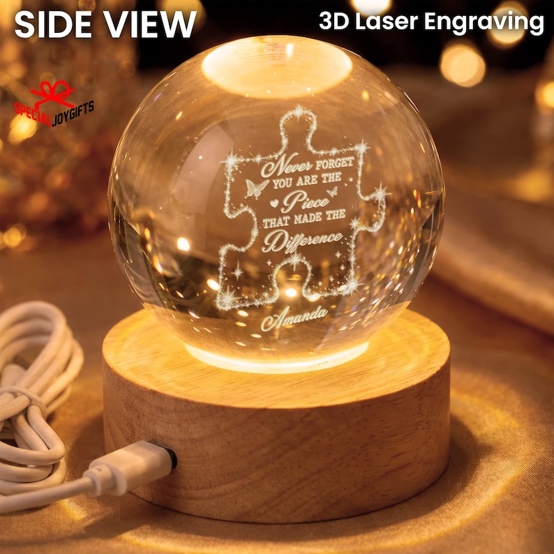 May include: A clear glass sphere with a 3D laser-engraved puzzle piece design featuring the text "Never forget you are the piece that made the difference" and the name "Amanda," resting on a round, light-colored wooden base with a white USB cord.