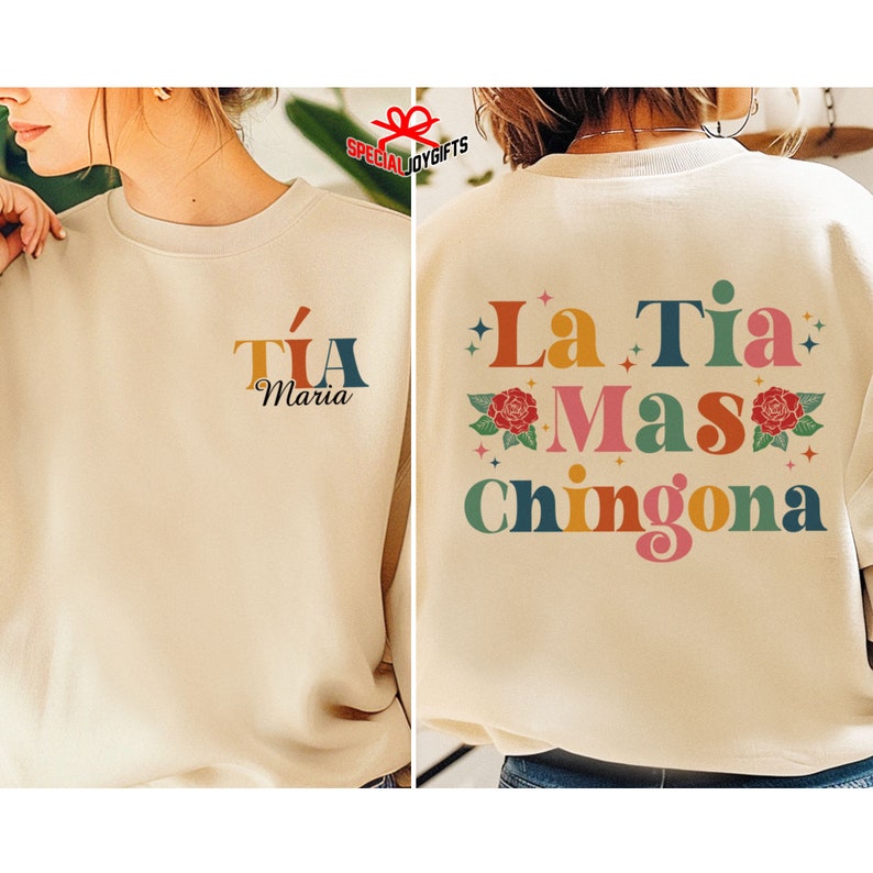 以下が含まれることがあります： ベージュのトレーナーで、背面には「La Tia Mas Chingona」というカラフルなグラフィックが描かれており、花と星が描かれています。トレーナーの前面には「Tia Maria」というカラフルなグラフィックが描かれています。