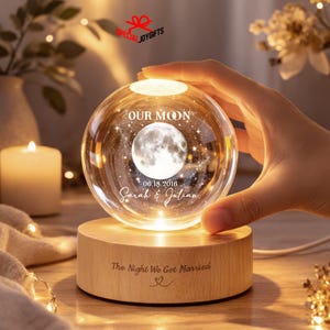 May include: A clear glass sphere lamp featuring an internal 3D laser-etched moon, the text "OUR MOON", "06.18.2016", and "Sarah & Julian". The sphere rests on a cylindrical light-colored wooden base engraved with "The Night We Got Married" and a heart.