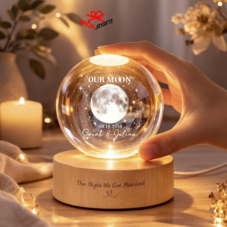 May include: A clear glass sphere lamp featuring an internal 3D laser-etched moon, the text "OUR MOON", "06.18.2016", and "Sarah & Julian". The sphere rests on a cylindrical light-colored wooden base engraved with "The Night We Got Married" and a heart.