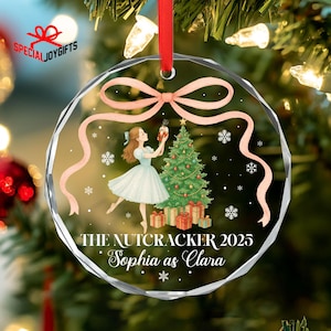 May include: A clear, round Christmas ornament with a ballet-themed design. The ornament features a ballerina, a Christmas tree, and the text "THE NUTCRACKER 2025 Sophia as Clara." A pink ribbon and snowflakes decorate the ornament.