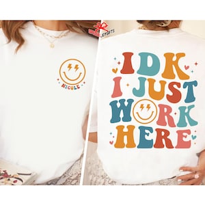 Pu&ograve; includere: Maglietta bianca con una grafica colorata che dice "IDK I JUST WORK HERE" con una faccina sorridente al centro della parola "WORK". La faccina sorridente ha due fulmini per gli occhi.
