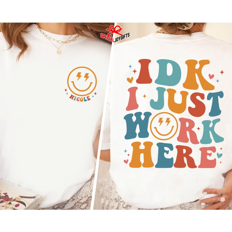 Pu&ograve; includere: Maglietta bianca con una grafica colorata che dice "IDK I JUST WORK HERE" con una faccina sorridente al centro della parola "WORK". La faccina sorridente ha due fulmini per gli occhi.