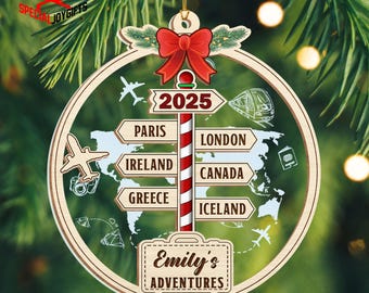 May include: Wooden Christmas ornament with a travel theme. The ornament features a red bow, a signpost with destinations like Paris and London, and the text "Emily's Adventures". The year 2025 is also displayed.